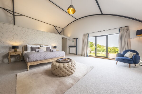A bedroom with a bed, bedside tables, a round ottoman, a blue armchair, and a large window with a view.