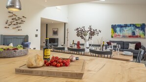 Een moderne eetruimte met een houten tafel, een snijplank met brood en tomaten, en een fles olijfolie. Er staan stoelen rond de tafel, een bank en een schilderij aan de muur.
