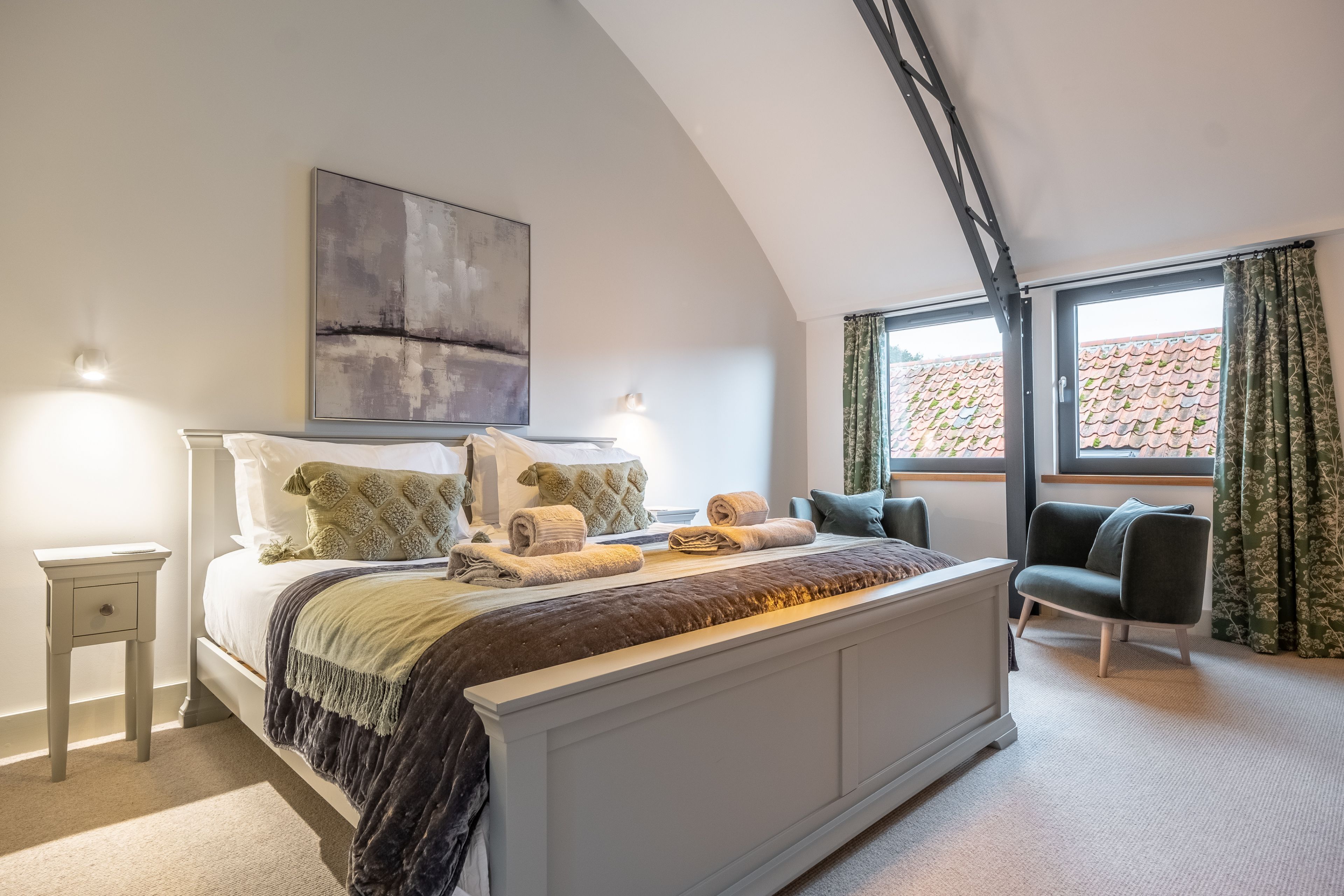 A bedroom with a large bed, a bedside table, a chair, a window with curtains, and a painting on the wall.