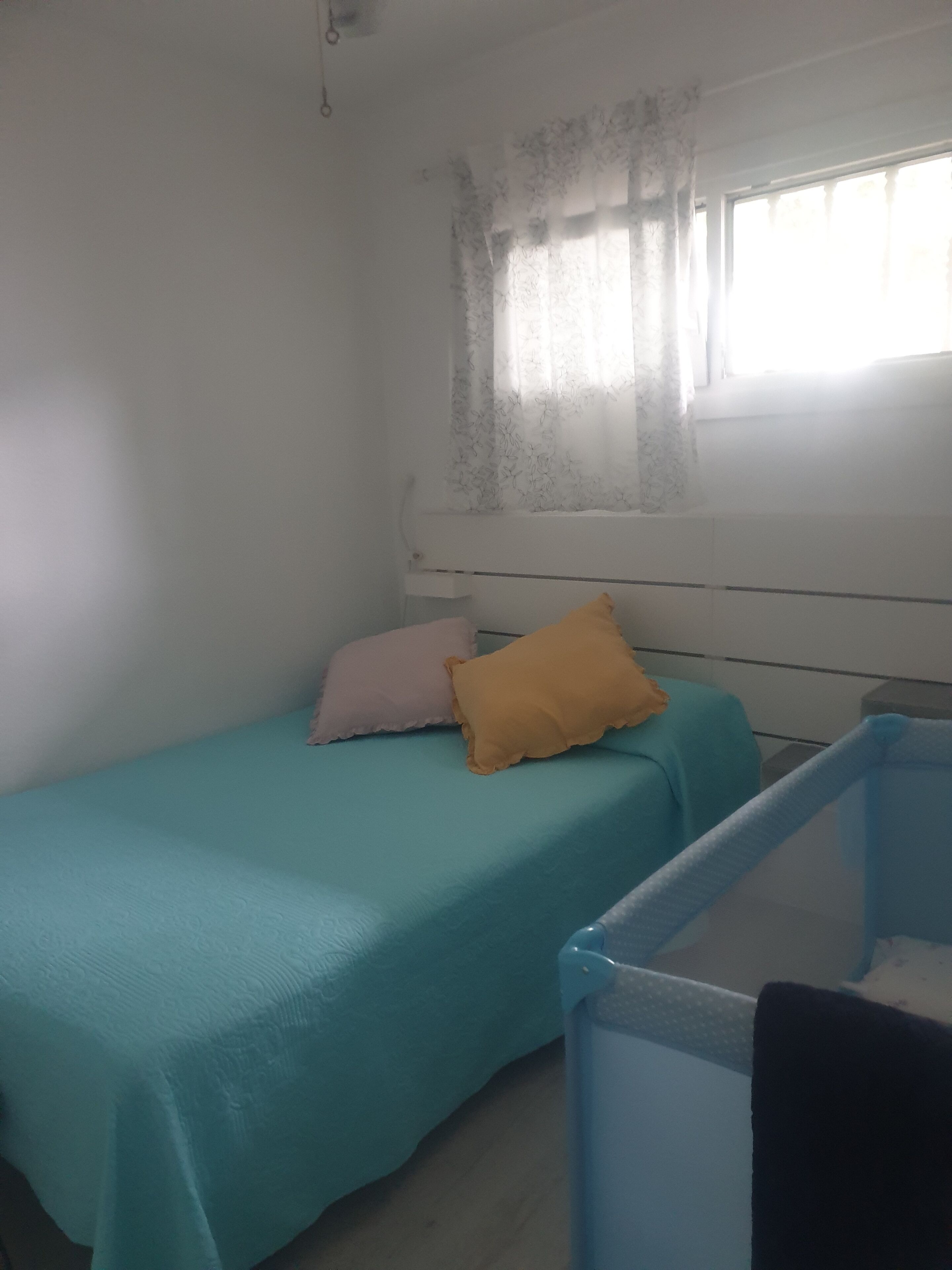 Una habitación pequeña con cama individual, colcha color esmeralda, dos almohadas, una ventana con cortinas translúcidas y una cuna con estructura azul.