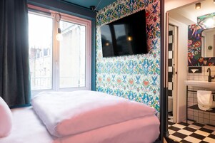 Un dormitorio con un ventanal grande, una cama con ropa de cama rosa, un televisor montado en una pared colorida y un baño con piso a cuadros y un lavamanos.