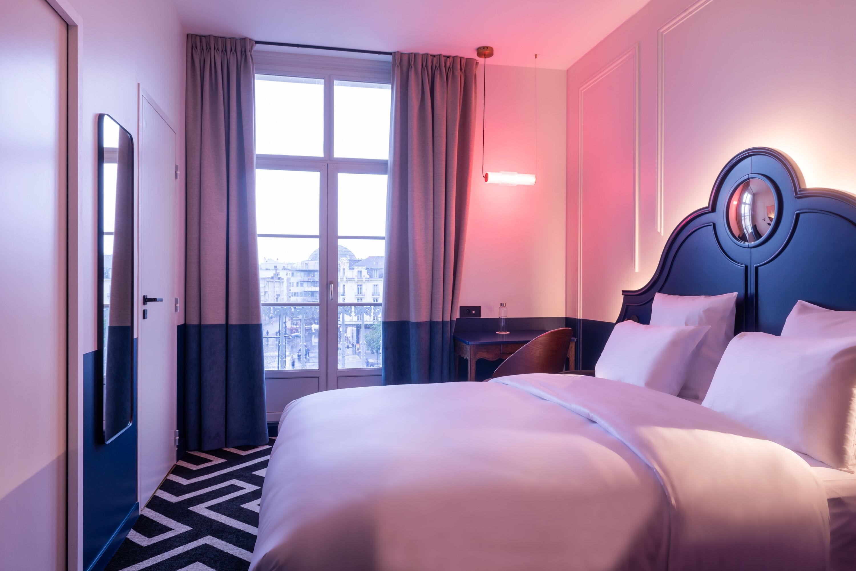 A hotel room with a large bed, a desk, a chair, and a view of the city through the window.