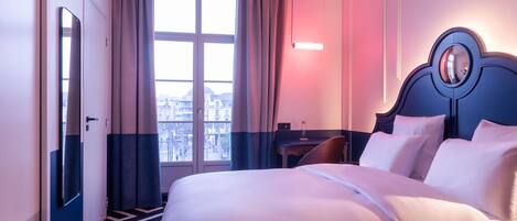 A hotel room with a large bed, a desk, a chair, and a view of the city through the window.