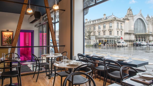 A restaurant with wooden floors, black chairs, and tables set for dining. Large windows offer a view of a historic building and a street with cars. - LABE Hôtel Tours Centre Gare (Tours)