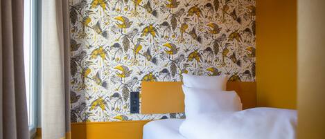 A hotel room with a bed, a nightstand, a wall-mounted lamp, and a window with curtains.
