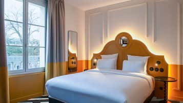A hotel room with a large bed, a window with curtains, a round mirror, and a small table.