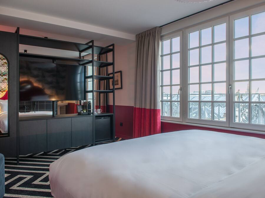 A hotel room with a large bed, a desk with a chair, a small table, and a large window with a view of a cityscape.
