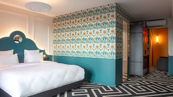 A hotel room with a large bed, a teal accent wall, patterned wallpaper, a black and white geometric carpet, and a small desk with a chair.