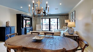 A dining area with a large wooden table, chairs, a chandelier, and a view of the outdoors through large windows. - Aegean #329 | Community Pool & Tennis + Shuffleboard (Destin)