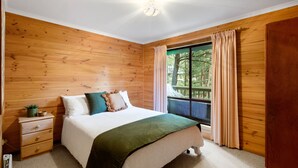 Une chambre avec un mur lambrissé en bois, un lit recouvert d’une couverture verte, une table de chevet ornée d’une plante et une fenêtre avec des rideaux.
