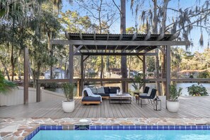 A covered outdoor seating area with a pool, surrounded by trees and a view of a lake.