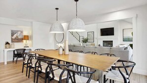 A modern dining room with a wooden table and black chairs, two hanging pendant lights, and a grey sofa in the background. - 8BR Lakefront Estate | Pool, Hot Tub & Lake Access (Savannah)
