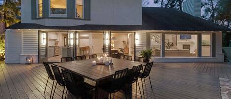 Una casa moderna con una amplia terraza, un comedor con mesa y sillas, y una cocina visible a través de puertas de vidrio.