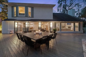A modern house with a large deck, a dining area with a table and chairs, and a kitchen visible through glass doors. - 8BR Lakefront Estate | Pool, Hot Tub & Lake Access (Savannah)