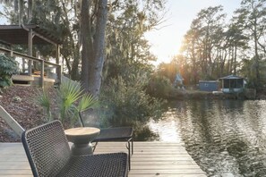 A wooden deck with outdoor seating, a calm body of water, and a forested area in the background.