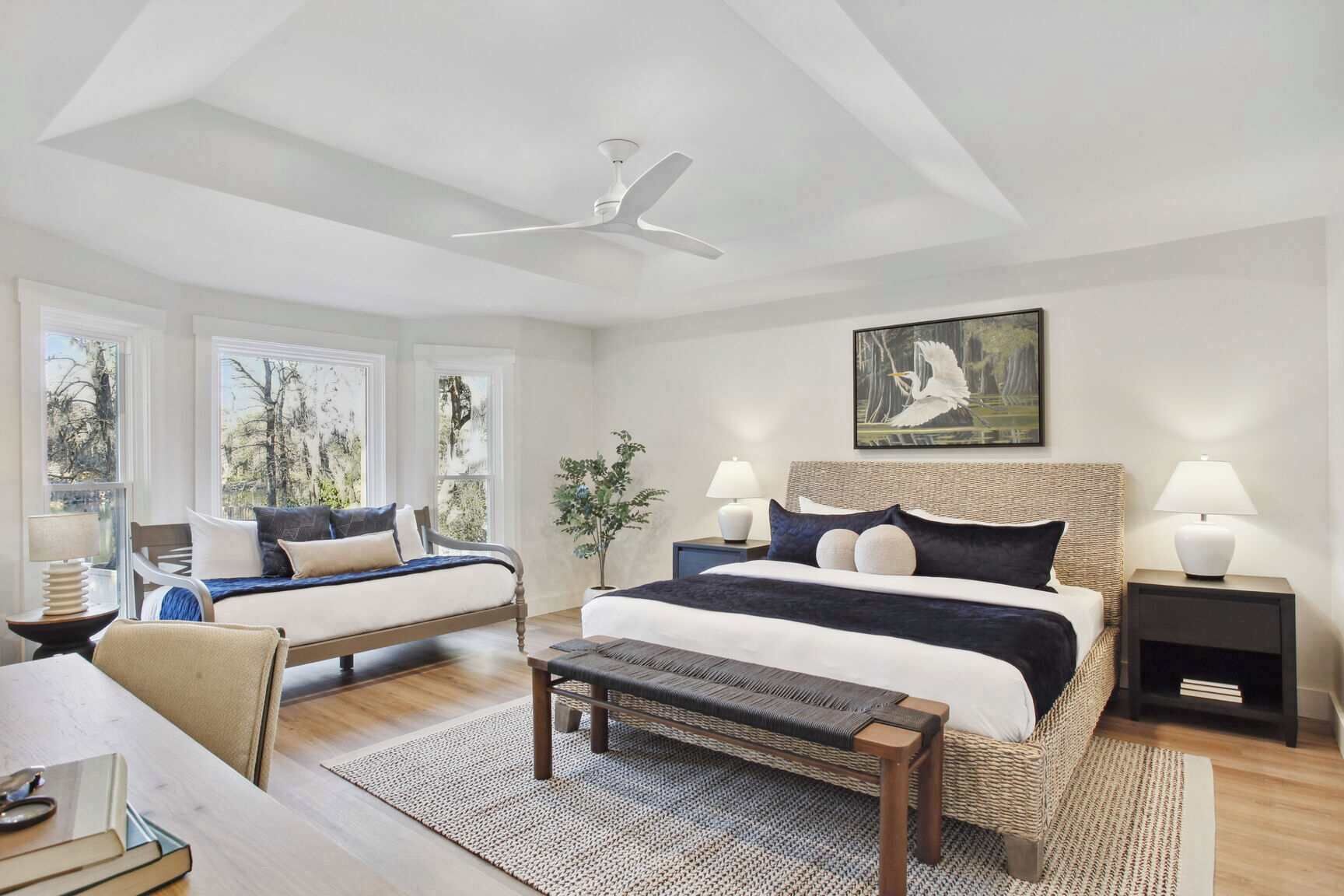 A modern bedroom with two beds, a ceiling fan, large windows, a painting, and a small table.