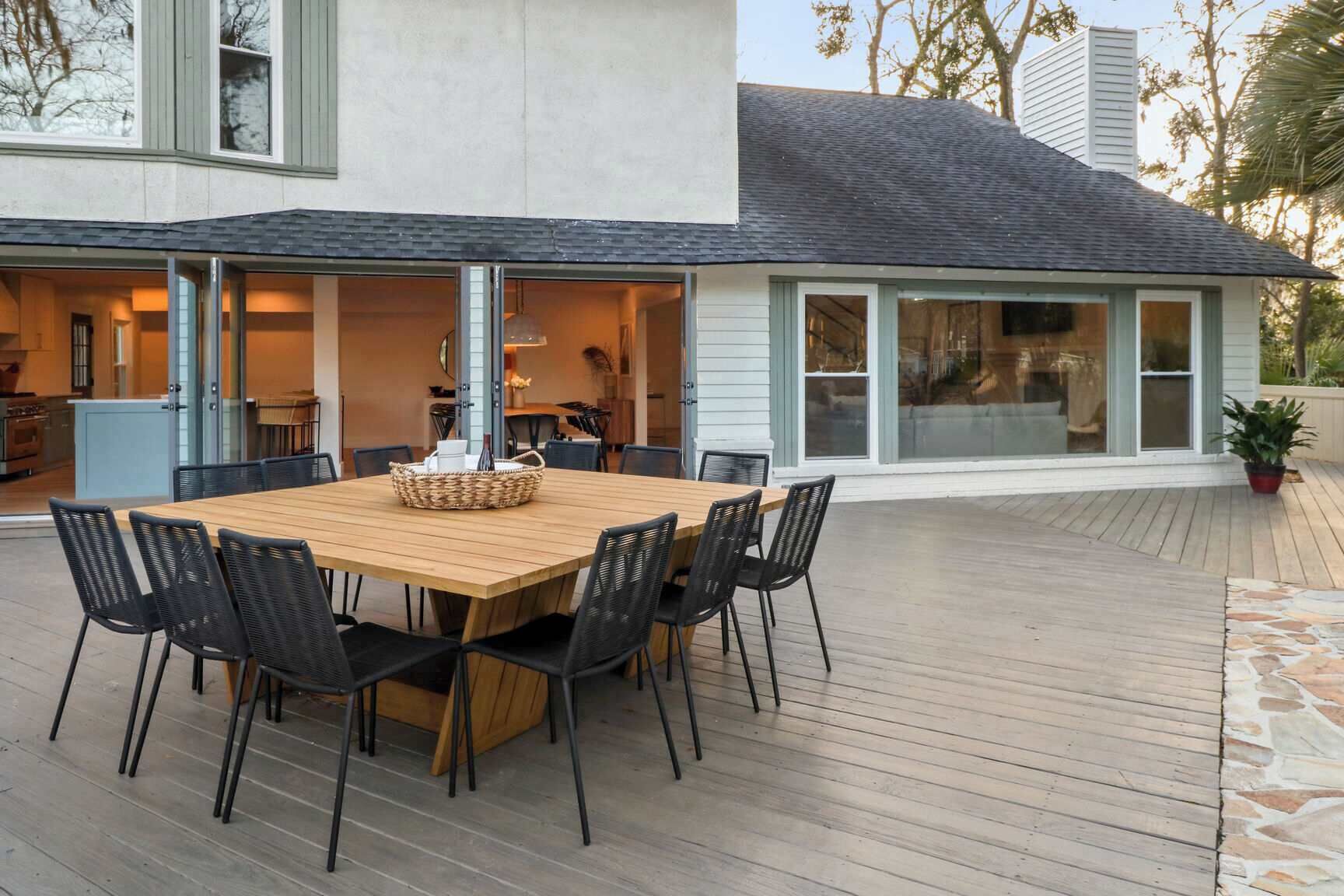 An outdoor dining area with a wooden table and black chairs, connected to a building with large windows and a stone pathway.