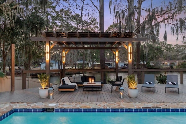 A poolside area with a wooden deck, seating, and a pergola with lights.