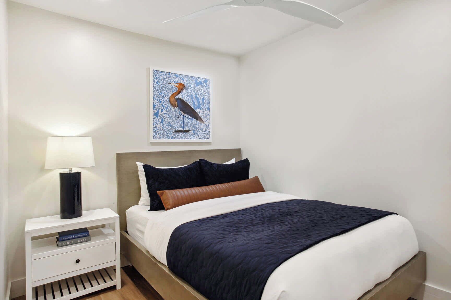 Un letto rifatto con cura, con una coperta blu scuro, un cuscino bianco e un cuscino marrone. Un comodino bianco con una lampada nera e un libro intitolato 'Kings Cages'. Sopra il letto, sulla parete, c'è un quadro con un uccello.