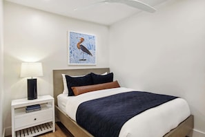 A neatly made bed with a dark blue bedspread, a white pillow, and a brown pillow. There is a white nightstand with a black lamp and a book titled 'Kings Cages.' A framed picture of a bird is on the wall above the bed. - 8BR Lakefront Estate | Pool, Hot Tub & Lake Access (Savannah)