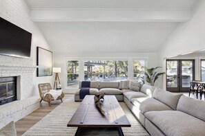 A spacious living room with a fireplace, a large sofa, a coffee table, and a view of a pool and outdoor seating. - 8BR Lakefront Estate | Pool, Hot Tub & Lake Access (Savannah)