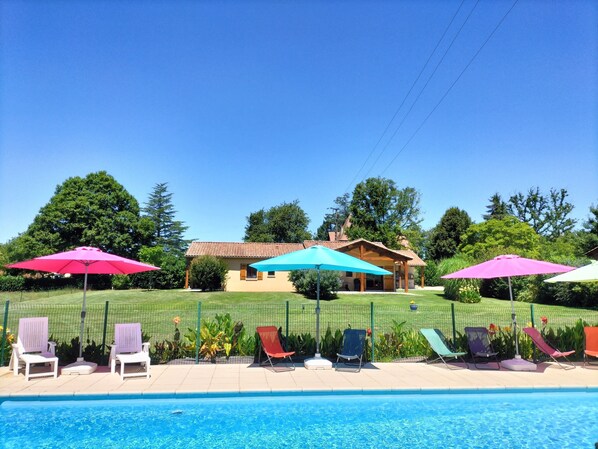 Una piscina con sillones de descanso y sombrillas, rodeada de una cerca y follaje.