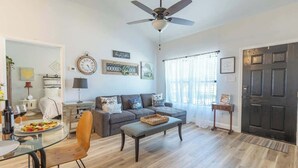 A living room with a sofa, a glass dining table, a ceiling fan, and a dark wooden door. - La Casita: Hot-tub & Firepit, 15 min to Fred! (Harper)