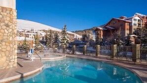 A pool area with a stone wall, a hot tub, and a view of snow-covered buildings and trees. - Grand Lodge Studio | Walk to Crested Butte Lifts, Pool, Hot Tub, Dog-Friendly (Crested Butte)