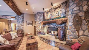 A stone fireplace with a log fire, a stone wall, a brown leather sofa, and a sign indicating 'SUITES'. - Grand Lodge Studio | Walk to Crested Butte Lifts, Pool, Hot Tub, Dog-Friendly (Crested Butte)