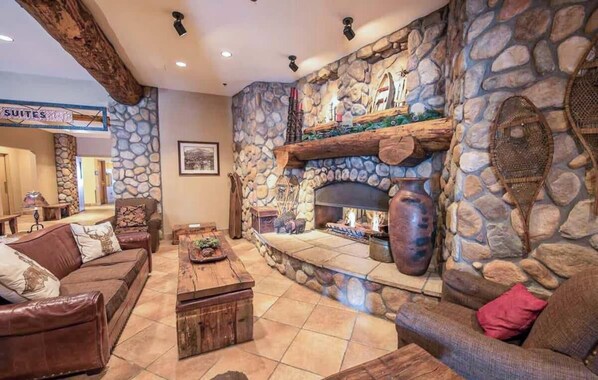 A stone fireplace with a log fire, a stone wall, a brown leather sofa, and a sign indicating 'SUITES'. - Grand Lodge Studio | Walk to Crested Butte Lifts, Pool, Hot Tub, Dog-Friendly (Crested Butte)