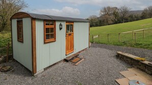 A small, light green metal shed with a wooden door and windows, situated on a gravel path with a stone wall and wooden fence in the background. - Rabbit Hill Hut (Abergavenny)