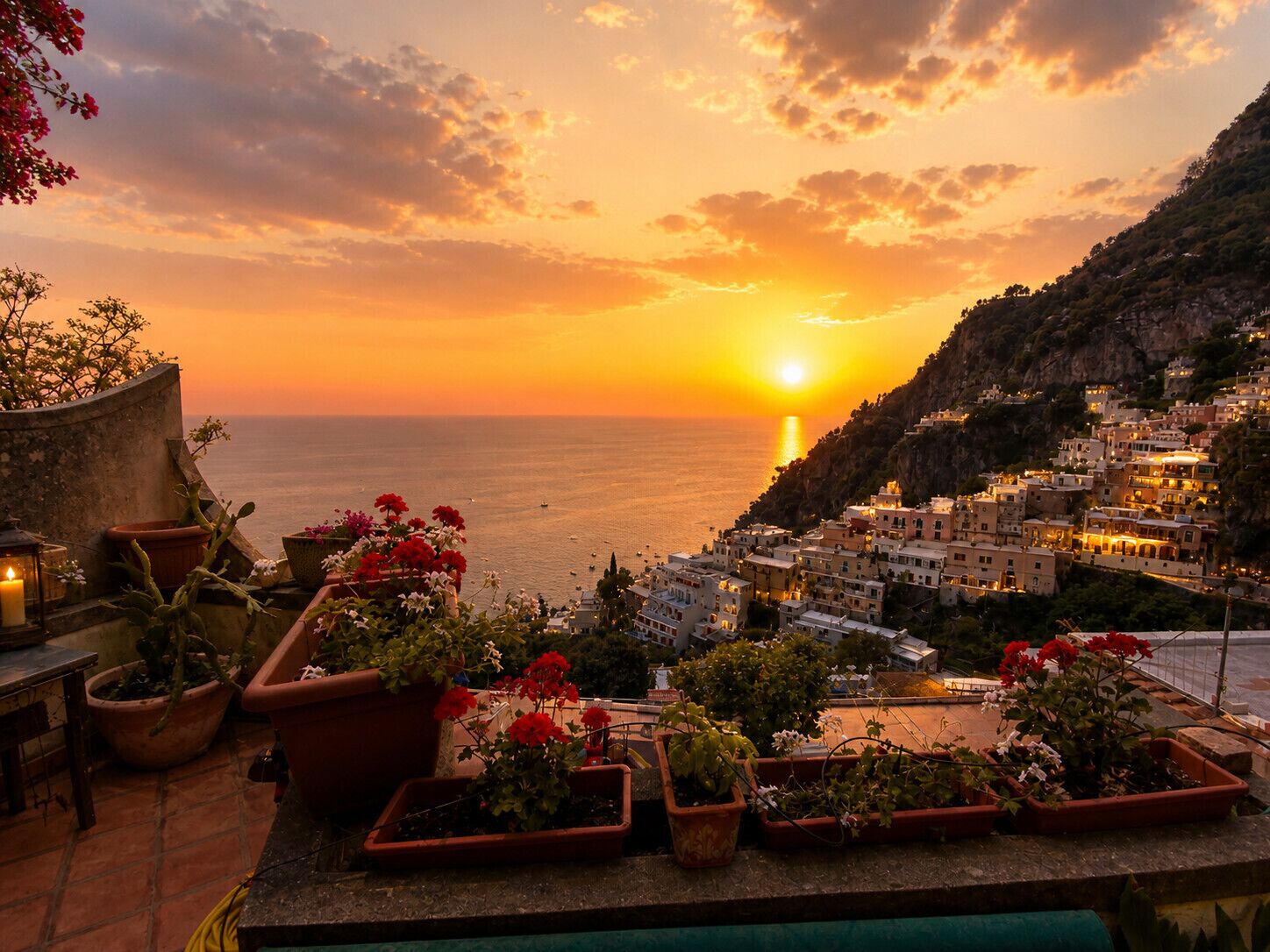 A coastal town at sunset with terraced houses, a view of the sea, and potted plants on a balcony.