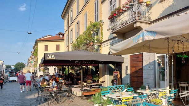 An outdoor café with tables and chairs, a sign for 'ADA', and a building with balconies.