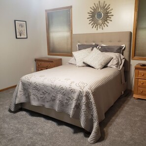 A bedroom with a bed, two nightstands, a window with blinds, a wall-mounted mirror, and a framed picture.