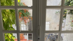 Une fenêtre avec un pot de fleurs sur le rebord, un vélo à l’extérieur, et une vue sur un jardin avec des fleurs oranges et des feuilles vertes.