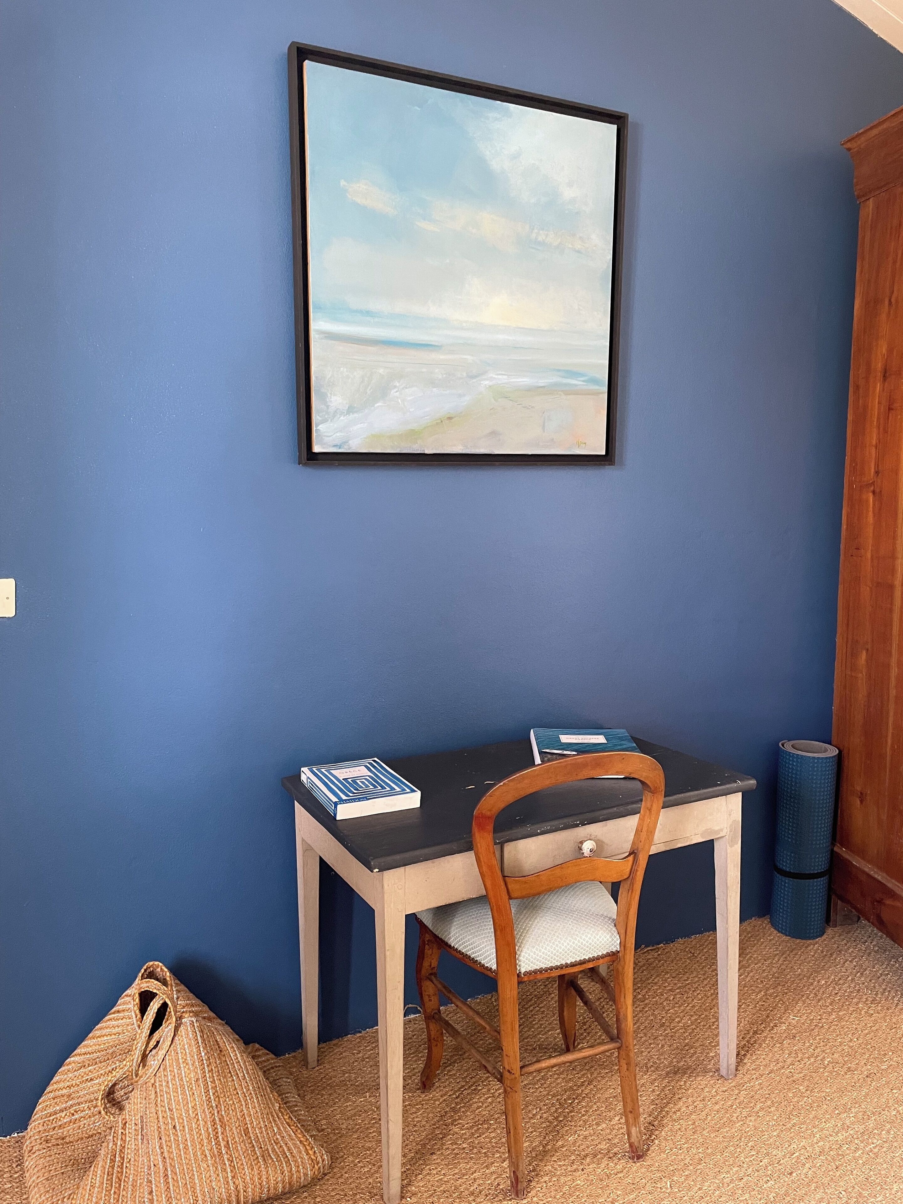 Une pièce avec un mur bleu, un tableau encadré, une chaise en bois, un bureau avec des livres et un sac en tissu tissé.