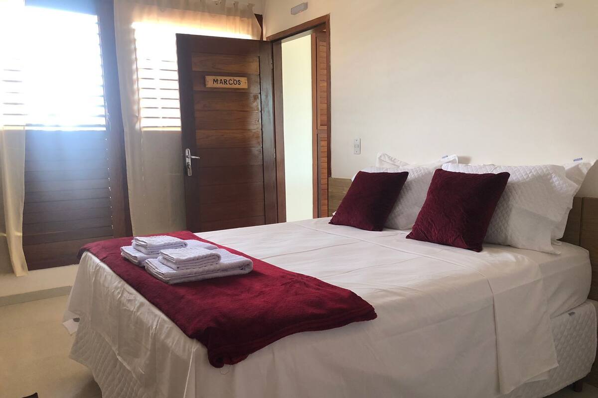 A neatly made bed with white and red bedding, a wooden door with a nameplate, and a window with blinds.