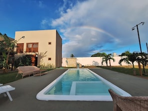 Piscina com espreguiçadeiras, um edifício com parede de pedra e uma palmeira.