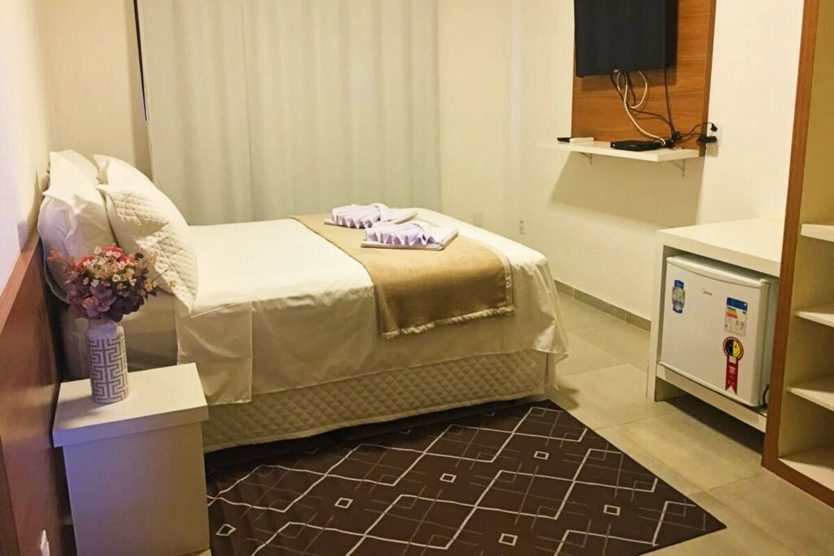 A hotel room with a bed, a small bedside table with a vase, a flat-screen TV mounted on the wall, and a mini-fridge.