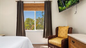 Une chambre à coucher avec un lit, un fauteuil en osier doté d’un coussin jaune, un téléviseur fixé au mur et une fenêtre avec des rideaux.