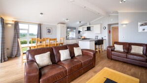 Een ruime woonkamer met een bruine leren bank en een houten salontafel, een eethoek met een houten tafel en stoelen, en een keuken met witte kasten en een centraal eiland.