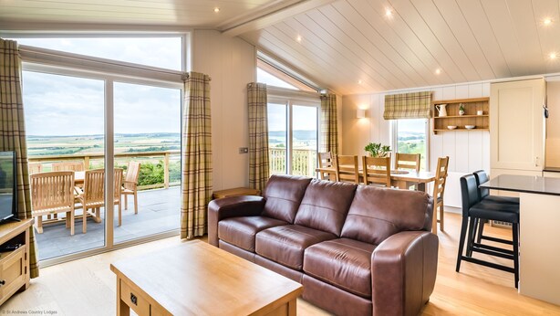 Een ruime woonkamer met een bruine leren bank, een houten eettafel en stoelen, een keuken met witte kasten, en een grote schuifpui die leidt naar een balkon met uitzicht op het platteland.