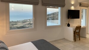 Un dormitorio con una cama, un escritorio y dos ventanas con vistas al mar y a las colinas.