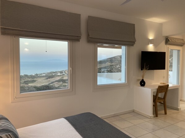 A bedroom with a bed, a desk, and two windows with views of the sea and hills.