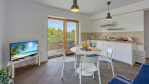 Une cuisine moderne avec des meubles blancs, un coin repas comprenant une table ronde et des chaises, un téléviseur posé sur un meuble, et un balcon offrant une vue sur les palmiers.