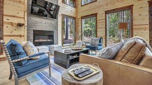A cozy cabin interior with a fireplace, wooden walls, and large windows offering a view of the forest. - Forever In Blue Jeans in Hochatown/Broken Bow,OK (Broken Bow)