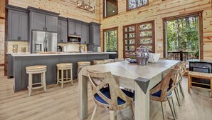 A spacious wooden cabin interior with a large kitchen, dining area, and multiple windows offering views of the surrounding forest. - Forever In Blue Jeans in Hochatown/Broken Bow,OK (Broken Bow)
