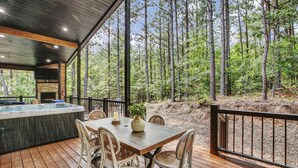 A wooden deck with a hot tub, dining table, and chairs, overlooking a forest. - Forever In Blue Jeans in Hochatown/Broken Bow,OK (Broken Bow)