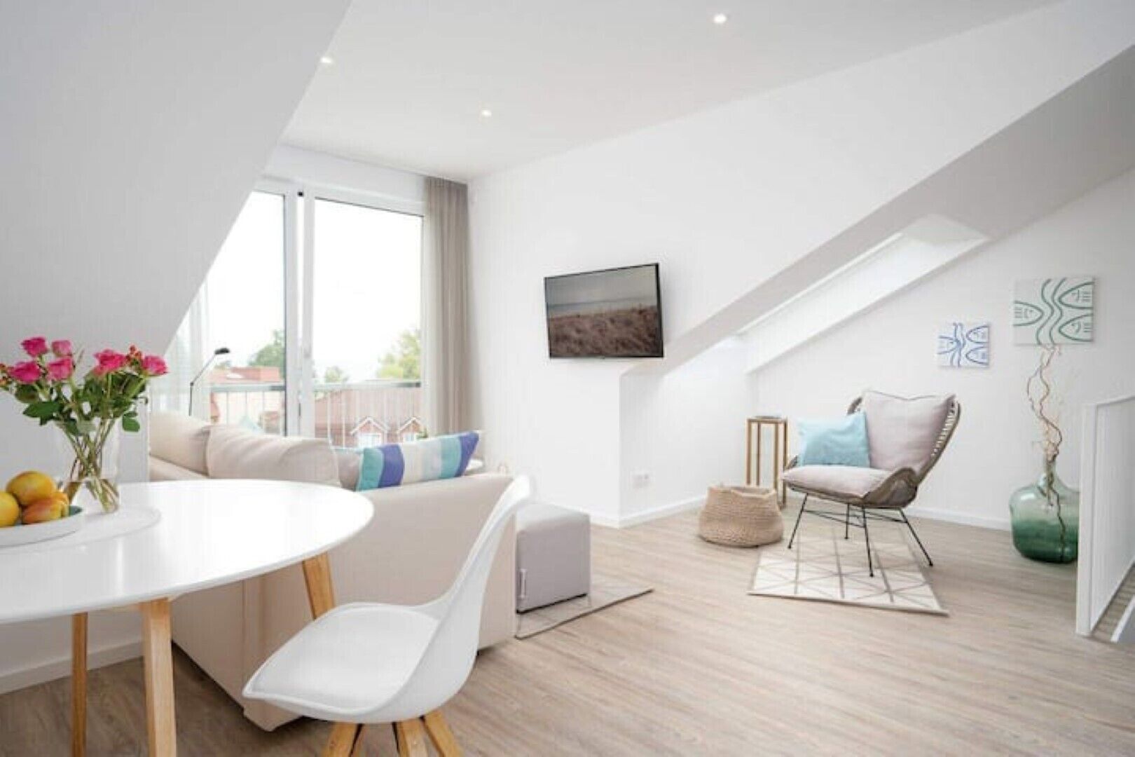 Ein modernes, gut beleuchtetes Zimmer mit einem weißen Sofa, einem Esstisch mit Blumenvase, einem Stuhl und Blick ins Freie durch ein großes Fenster.