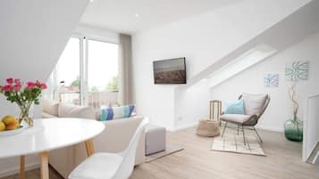 Ein modernes, gut beleuchtetes Zimmer mit einem weißen Sofa, einem Esstisch mit Blumenvase, einem Stuhl und Blick ins Freie durch ein großes Fenster.